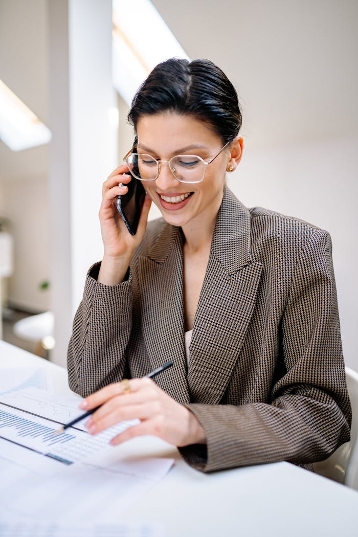 services-01 Confident businesswoman wearing glasses, talking on phone while reviewing documents in modern office setting.